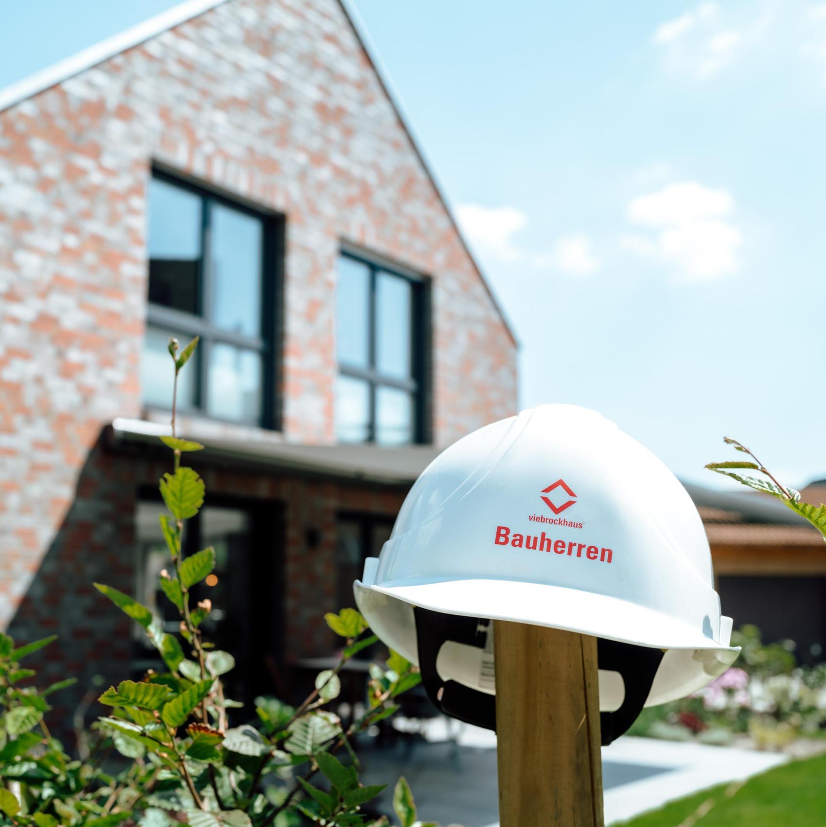 Ein Bauhelm hängt vor einem Neubau an einem Holzpfosten